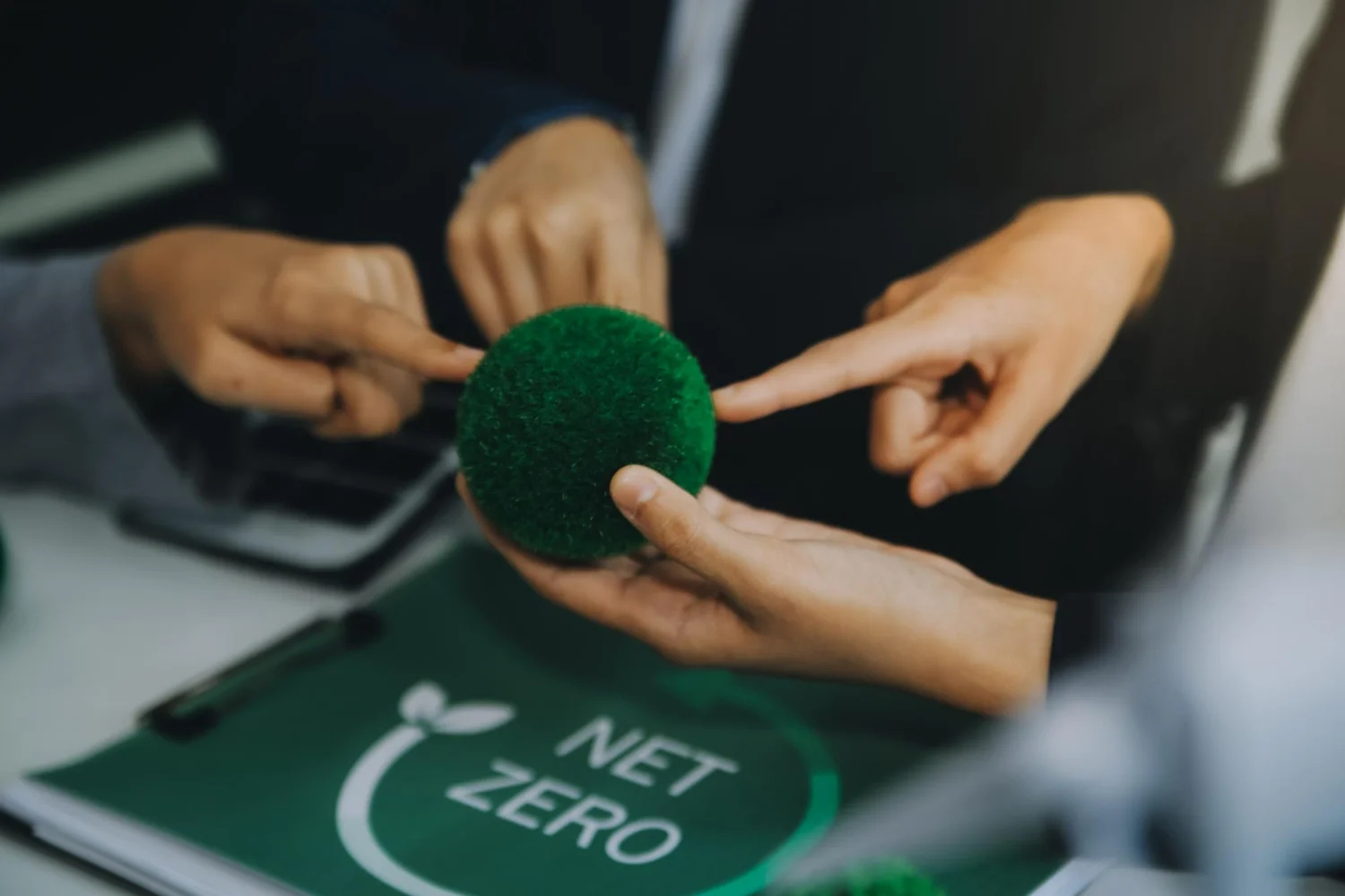 algumas mãos apontando e segurando uma pequena esfera verde e ao fundo um documento verde escrito net zero