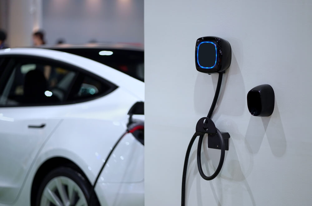 ao lado direito um carro elétrico da cor branca e ao lado esquerdo um carregador elétrico disponível para uso
